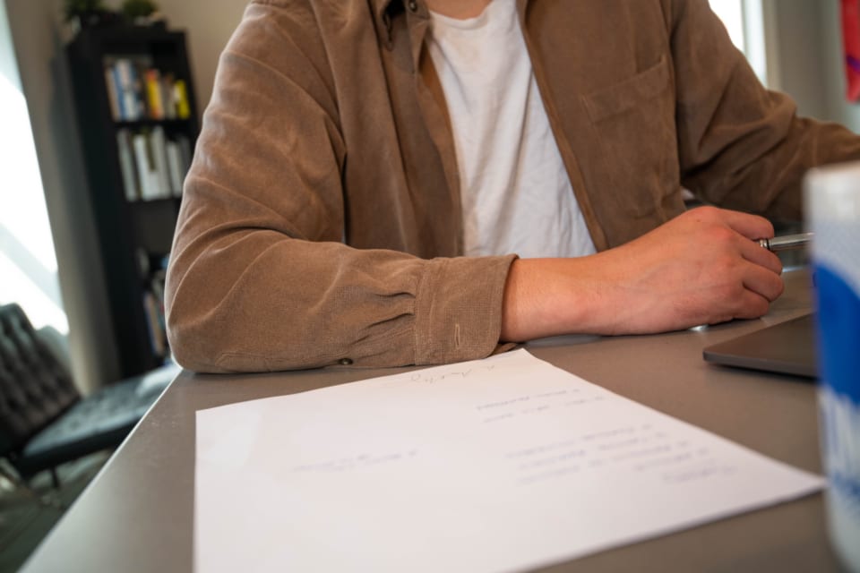 Une personne assise à une table, un stylo à la main, devant une feuille posée à côté d'un ordinateur portable.