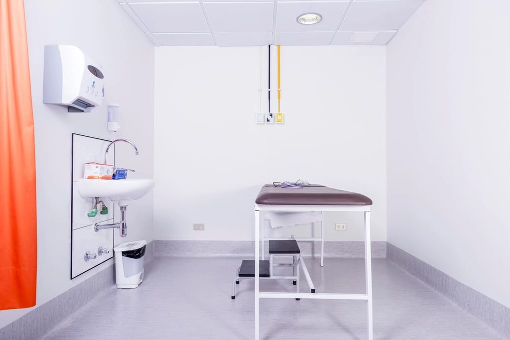 Une salle d'examen médical vide, avec une table d'examen, un marchepied et un lavabo, sous une lumière blanche.