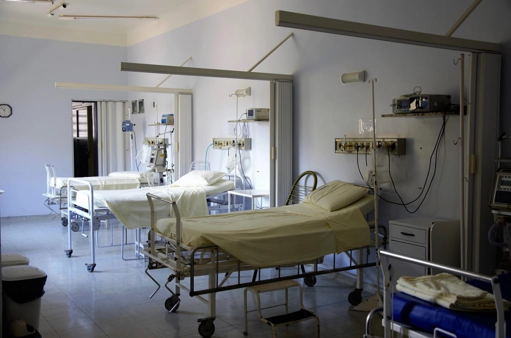 Une chambre d'hôpital commune et vide : plusieurs lits médicalisés aux draps blancs, des rideaux de séparation et des prises murales de fluides médicaux.